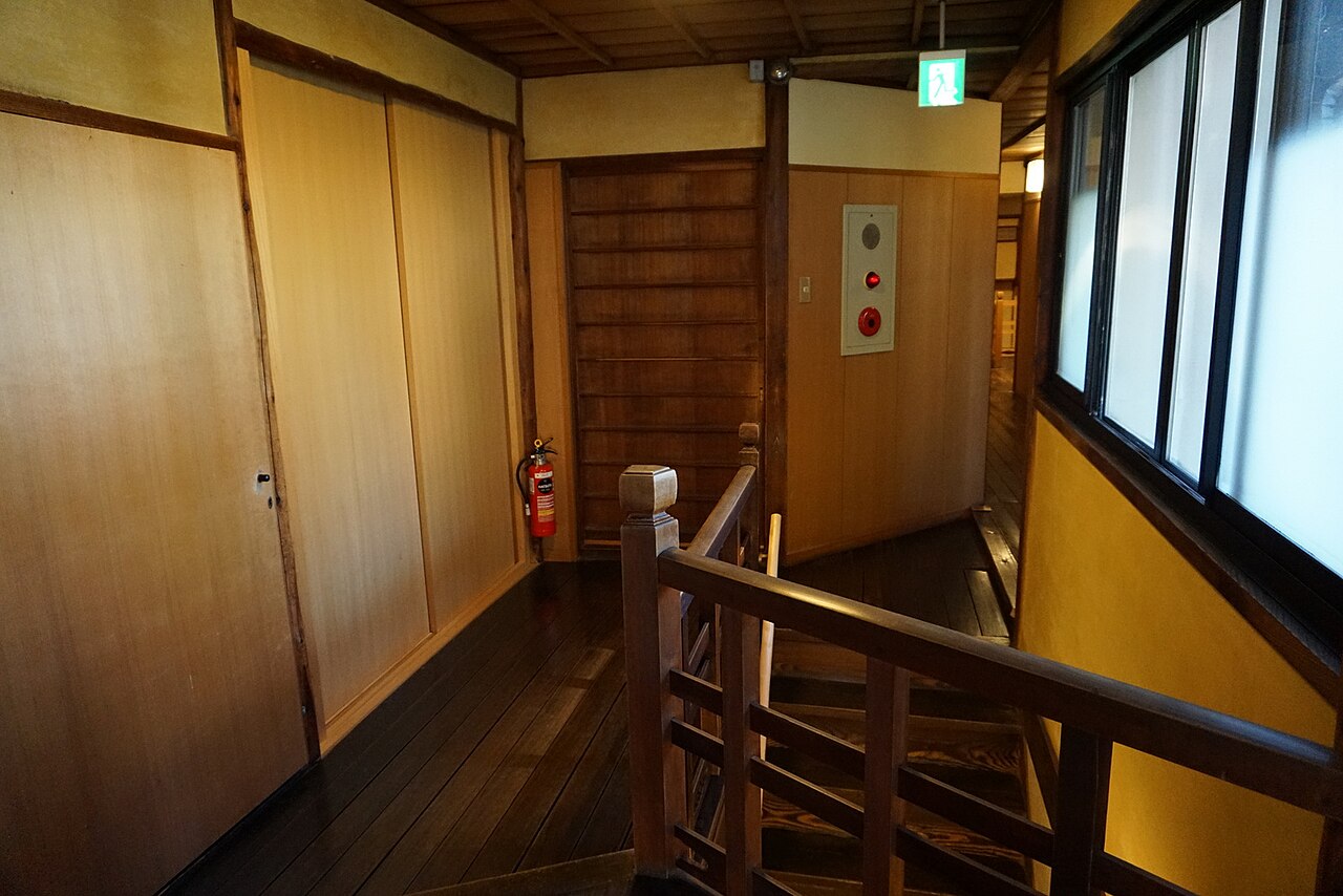 The upper-floor landing of a traditional Japanese inn: a dark polished wooden floor, a wooden balustrade around the stairwell, sliding doors along the wall, and low light.