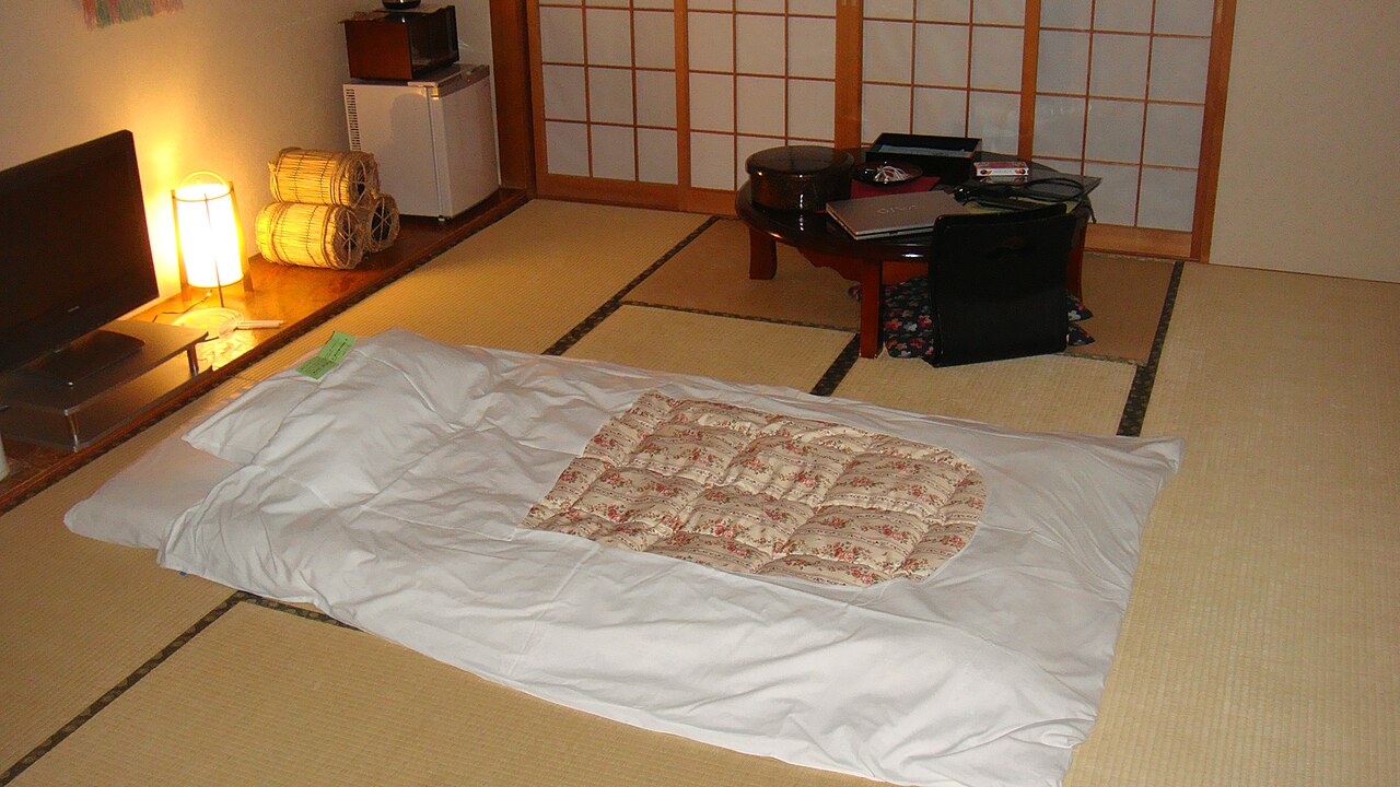 A ryokan guest room at night: a futon laid directly on the tatami floor, a low table with a legless floor chair behind it, and low lighting.